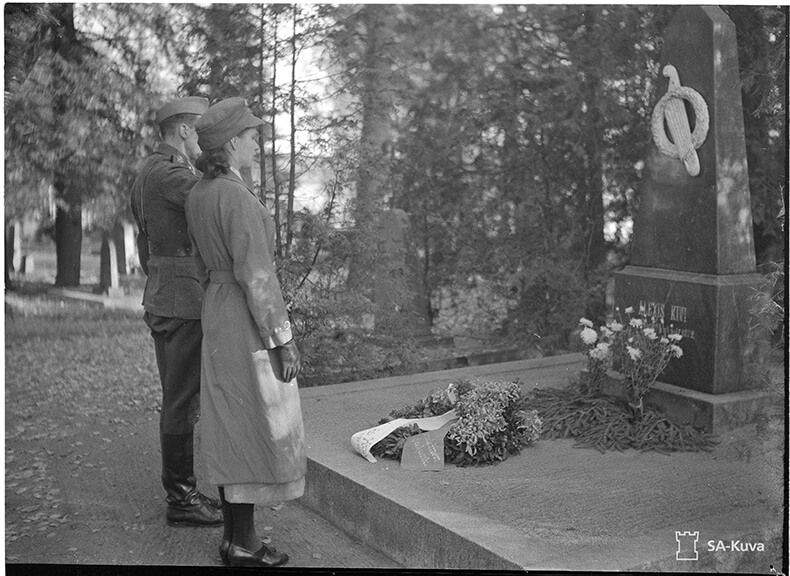 Sotilas ja lotta seisomassa Aleksis Kiven haudan edustalla, haudalla seppeleitä.