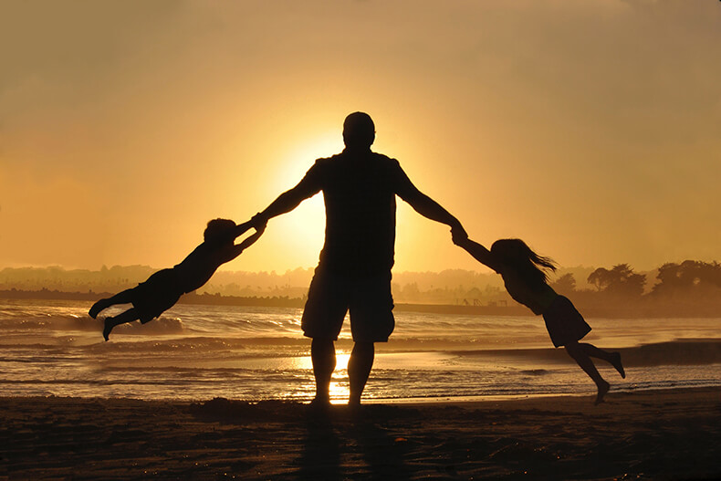 Isä pitää lapsia käsistä kiinni ja nostaa ilmaan, taustalla meri ja auringonlasku.