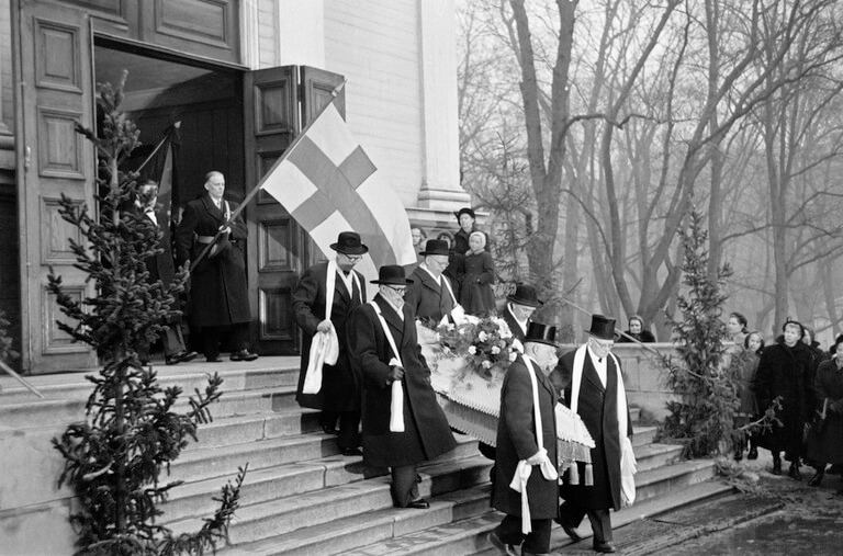 Miina Sillanpään hautajaissaattue kävelee kirkon portaita alas arkkua kantaen, kuvan sivulla näkyy Suomen lippu.