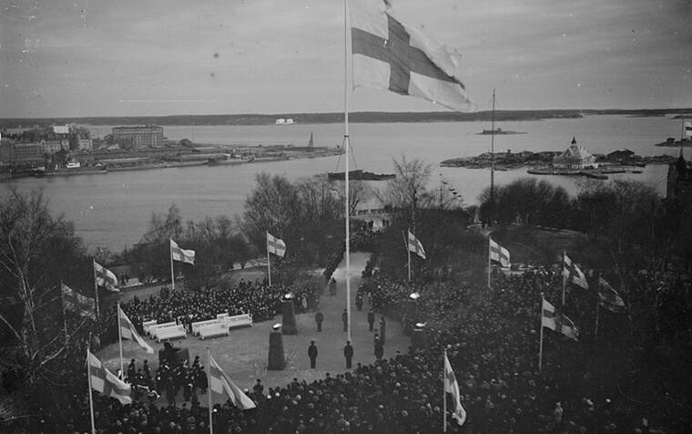 Mustavalkoinen maisemakuva Helsingin Tähtitorninmäeltä, Suomen lippu liehuu salossa ja taustalla näkyy meri. Suuri joukko ihmisiä on kokoontunut lipputangon ympärille.