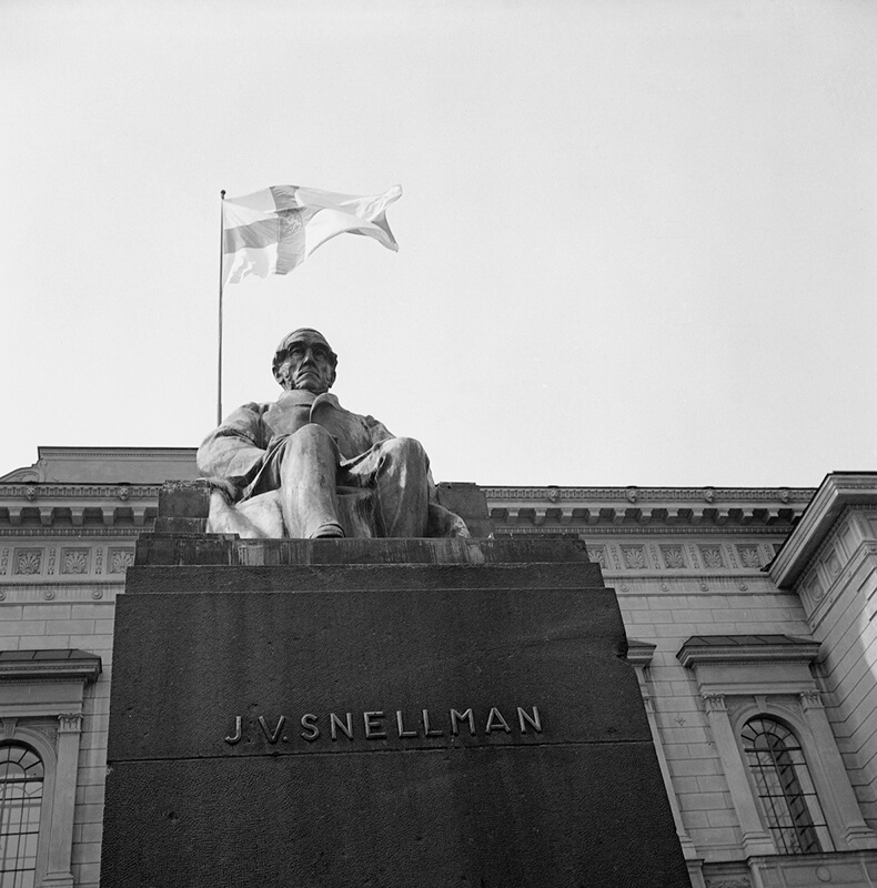 Johan Vilhelm Snellmanin patsas Suomen Pankin edustalla, taustalla Suomen lippu.