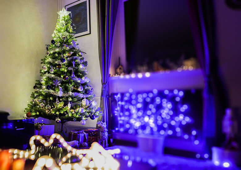 Huoneen nurkassa valkoisin ja hopeisin koristeltu joulukuusi, latvassa tähti. Sivulla sinisen sävyisiä jouluvaloja.