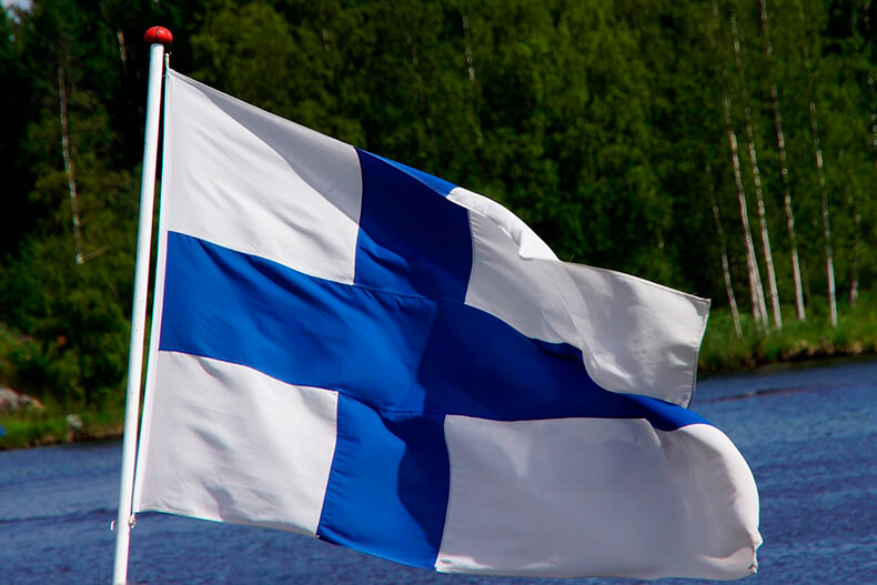 Suomen lippu liehuu tuulessa, taustalla vettä ja metsää.