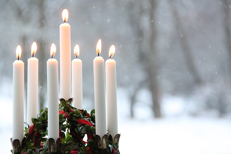Seitsemän kynttilää palamassa jouluisen koristeen päällä, taustalla luminen maisema.