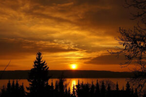 Kullanvärinen auringonlasku järvi- ja metsämaiseman yllä.