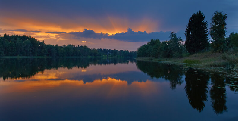Auringonlaskun järvimaiseman yllä, horisontista kajastaa hämäränsäteitä.