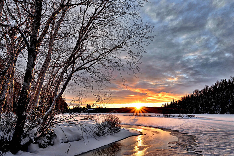 Keväinen järvimaisema, jää on jo osittain sulanut ja aurinko laskee värikkäästi horisontin taakse.