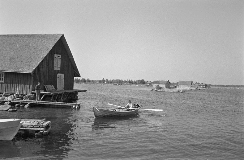 Mustavalkoinen kuva. Mies soutamassa venettä. Toinen mies seisoo laiturilla lähellä rannalla olevaa rakennusta.