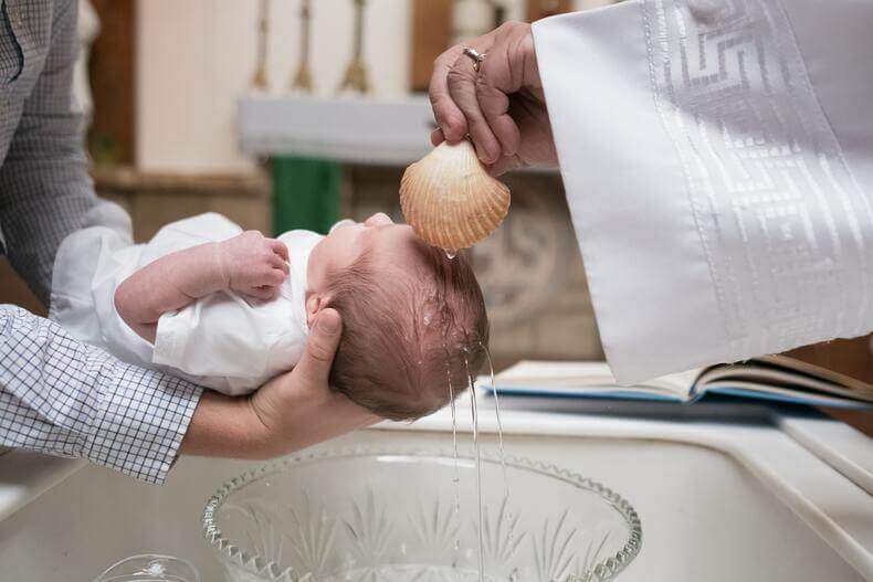 Pienen vauvan kastetilaisuus. Pappi kaataa vettä vauvan pään päälle isän pitäessä lasta kastemaljan yllä sylissään.